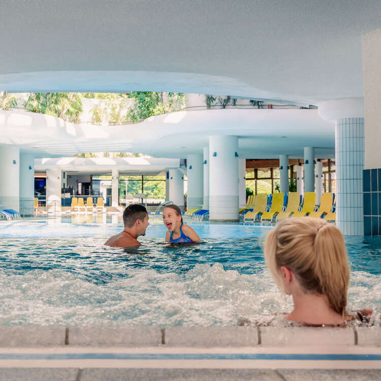 Mutter genießt die Massagedüsen im Warmwasserbecken und sieht ihrer Tochterund dem Papa beim Planschen zu